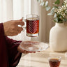 Person holding a glass mug with a leaf design, filled with a dark liquid, on a light-colored surface.